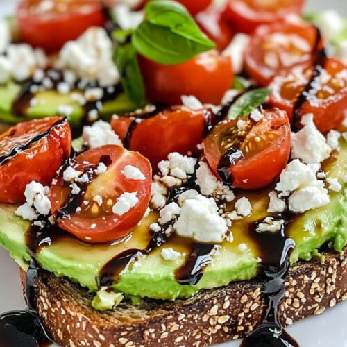 Avocado toast topped with cherry tomatoes, feta cheese, and balsamic glaze on a white plate